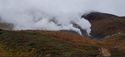 A geothermal landscape in Japan, showing steam rising from fumaroles on a rocky, barren mountainside. The foreground features patches of green and reddish vegetation, while thick clouds and mist partially obscure the background. A narrow trail winds through the terrain, leading toward the steaming vents, indicating volcanic activity typical of Japan's mountainous regions.